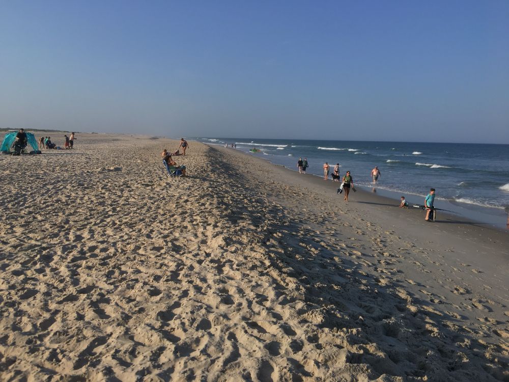 A photograph looking along a beach with the ocean to the right. The sand in the foreground is tracked with many footprints. A handful of figures with various beach gear are scattered in the mid ground. A low line of vegetation marks the top of the dunes on the left.