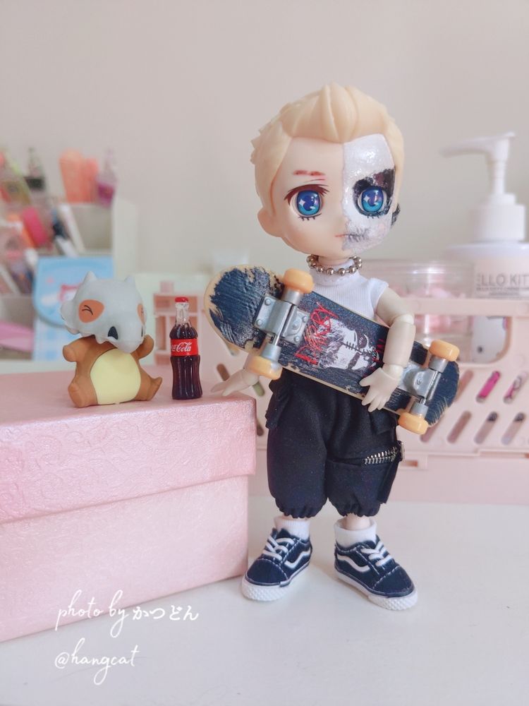 my custom ob11 doll, resembling darby allin. the doll is holding a skateboard, wearing a white tank top, cargo pants and vans sneakers. next to him there's a mini coke and a karakara pokemon.