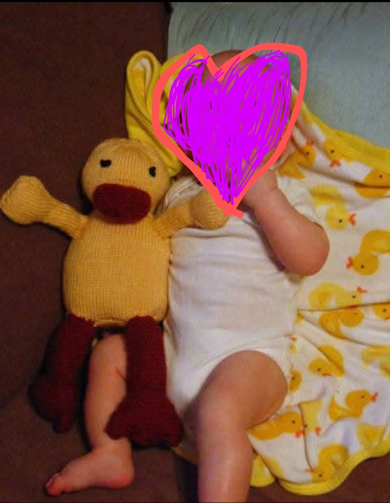 Baby with a knitted duck with a baby blanket with printed ducks.  Babys face has been scribbled over with a heart