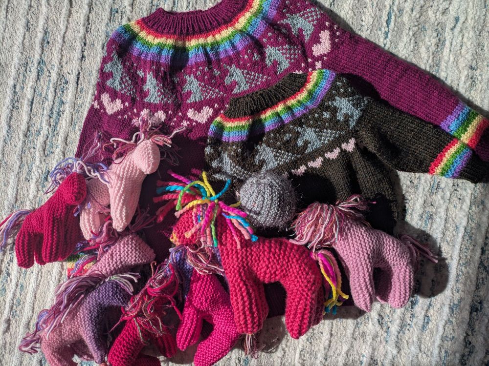 Against a white carpet are two handknitted sweaters with rainbow, unicorn and heart yoke and rainbow details on the sleeve.  The larger one is dark pink and the smaller is brown.  8 handknitted pink ponies in assorted shades of pink are scattered on top of the sweaters.