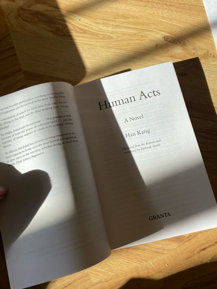 a book on a wooden table open to the inner title page. the sun is casting heavy shadows on the white pages. the book is human acts by han kang. 