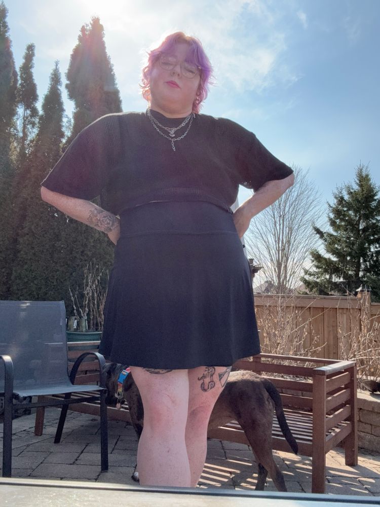 a fat white person with short purple hair stands in a back yard showing their black mess top and a short black skirt. 