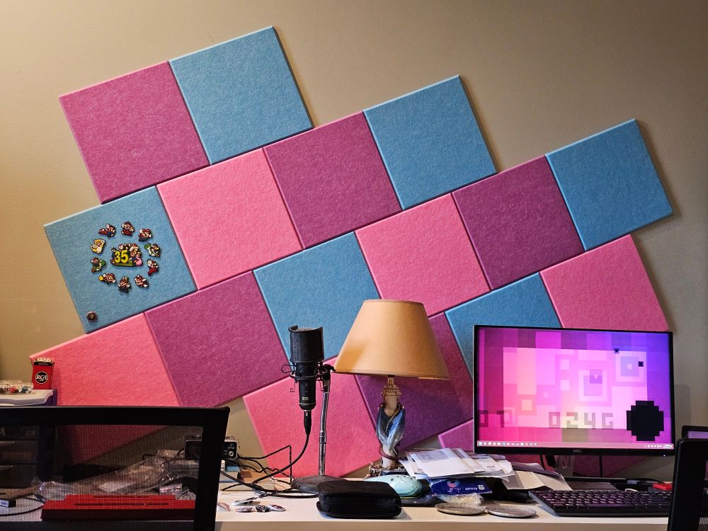 On the wall behind a cluttered desk is a series of square corkboard tiles, arranged diagonally in a repeating pattern of blue, purple, and pink tiles.