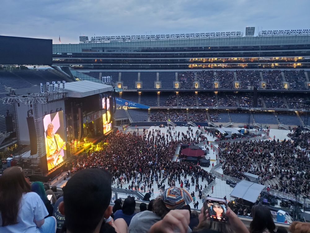 Inside Chicago Soldier Field from up high, looking down at the standing crowd in front of a stage. A man in yellow is displayed on the screen, playing on a guitar, with the shirt saying "DEVO".