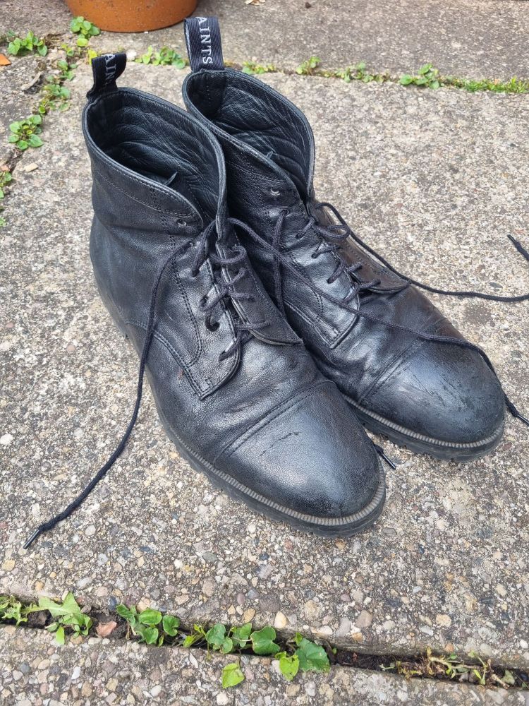 A pair of mildly worn black boots with laces, set on a paving stone outside.