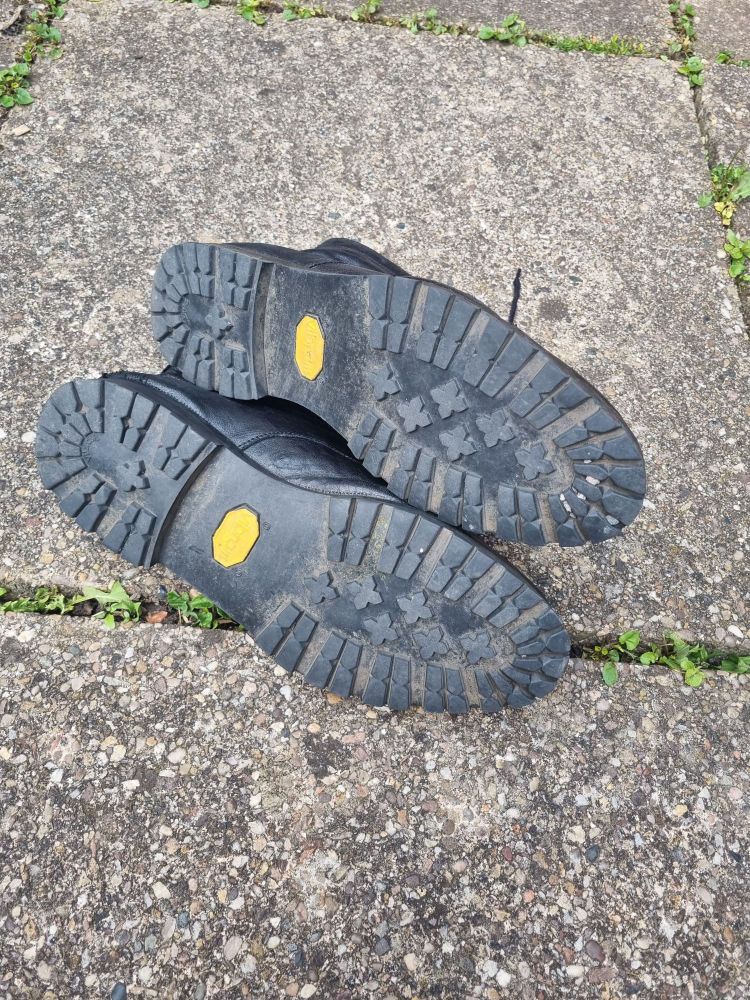 The same pair of mildly worn black boots on a paving stone outside, but now turned over so we can see the luscious new soles, stronger and heartier than ever.