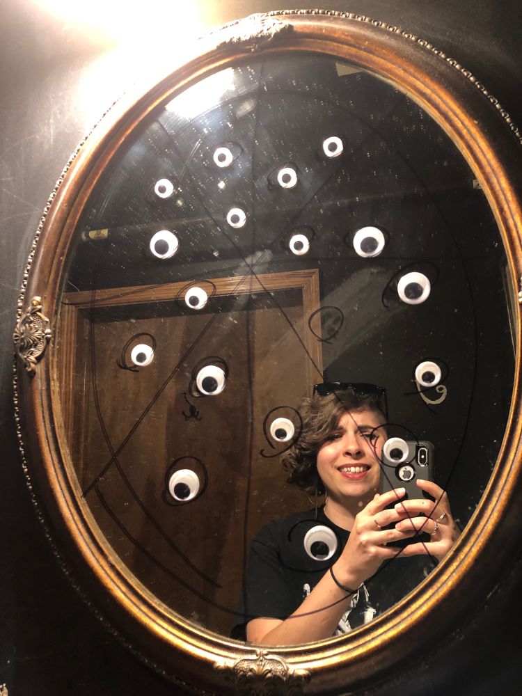 An amused person takes a selfie in a mirror. That mirror is vandalized with sharpie to make a pepperoni pizza. The “pepperonis” now have googley eyes on them.