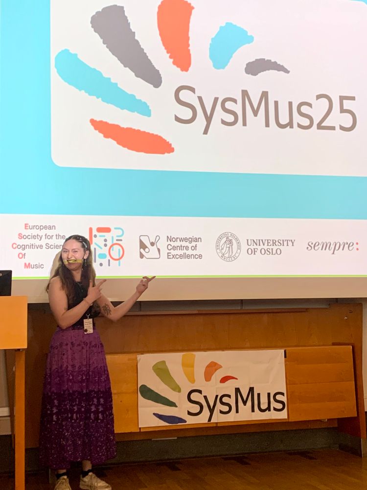 Hazel standing in front of and pointing at the SysMus logos before her spoken presentation