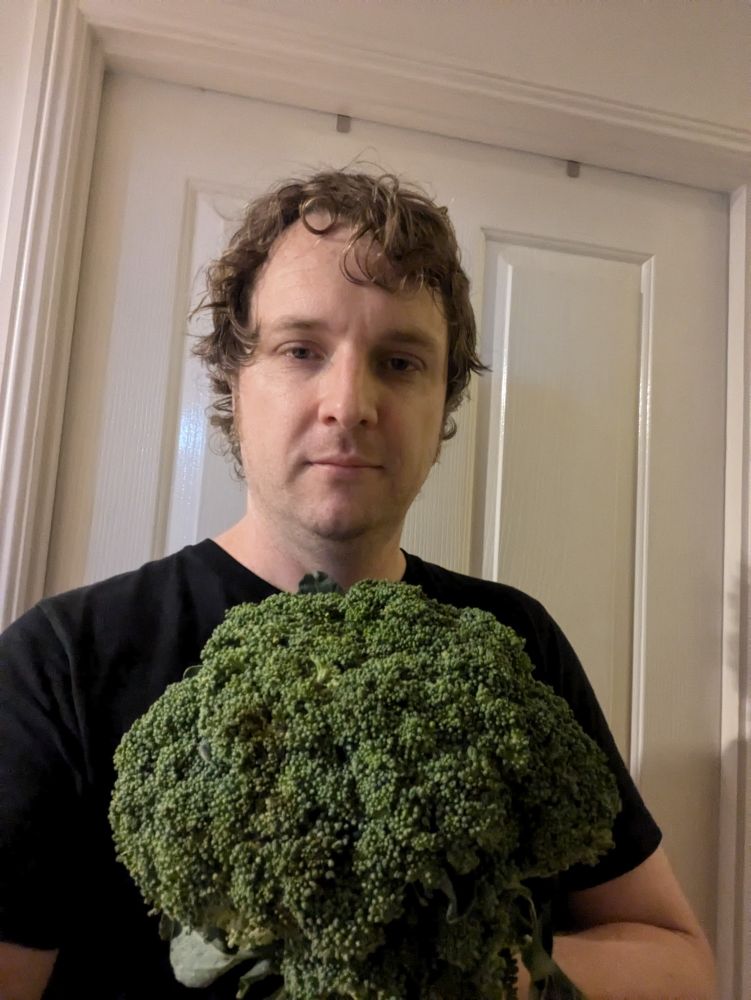 A sweaty man, possibly manic, holding a gigantic head of broccoli 