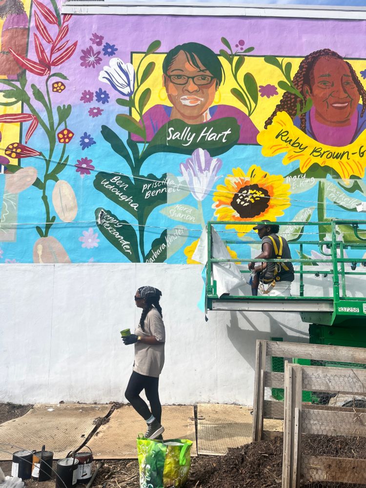 A progress shot of two artists, Brittany Baum and De’von Downes, helping to paint and install the urban farm mural