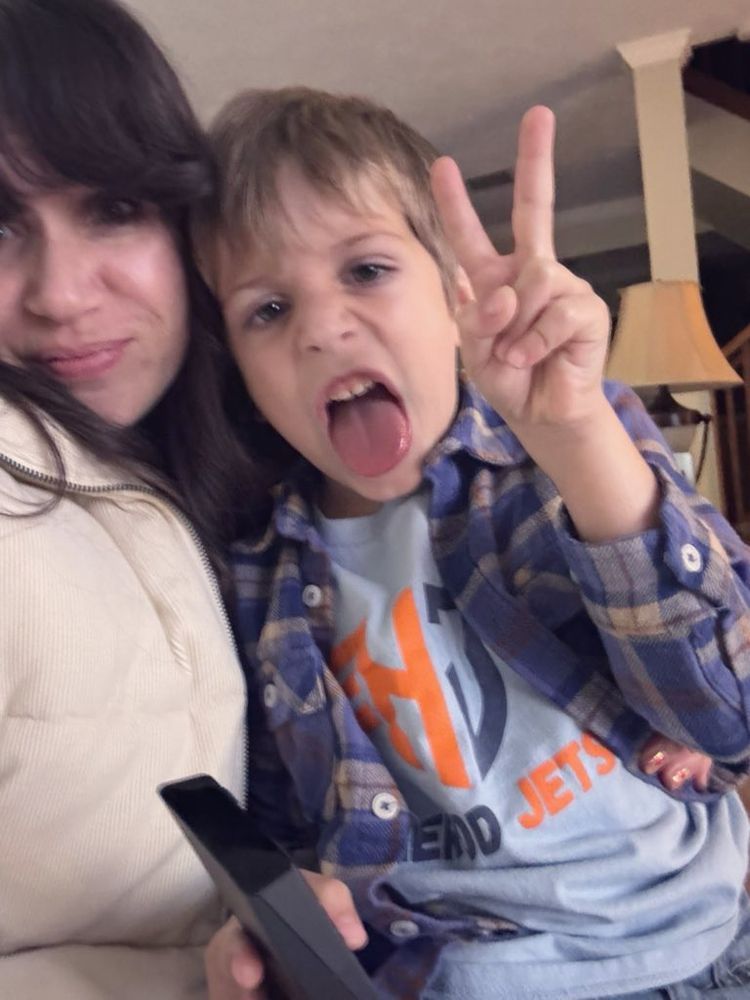 Young boy sitting with his mom making a silly face sticking his tongue out and holding up a peace sign 