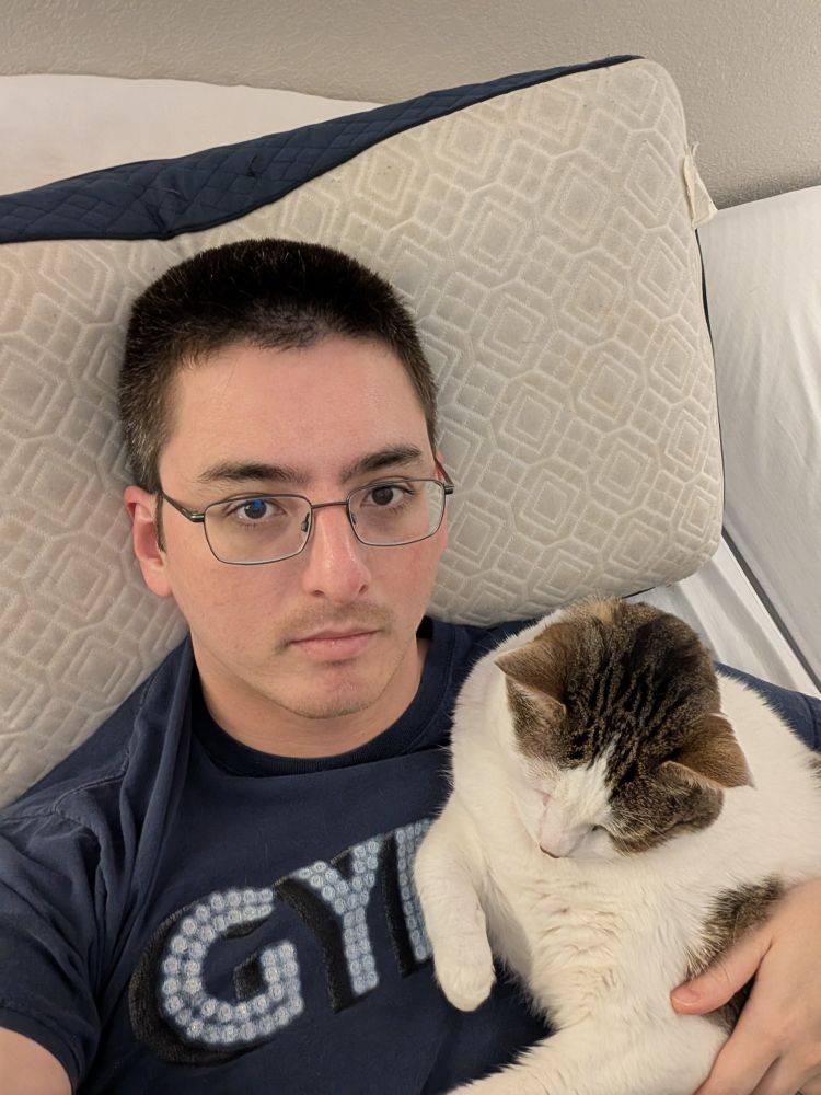 Me on bed in a shirt from the Patti LuPone production of Gypsy from 2008, also my big ass cat Carl is cuddling with me as per his usual. Also also I'm washing my bedsheets so my bed is all stripped down 