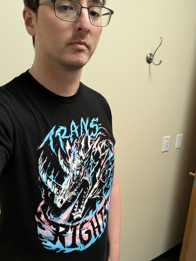 Selfie of me wearing a black graphic T-shirt shirt that says "Trans Rights", but the text is above and below a badass demon-dragon thing. Besides black the only other colors are white, pink, and light blue, aka the trans flag colors. The font and design look like they belong on a heavy metal album.