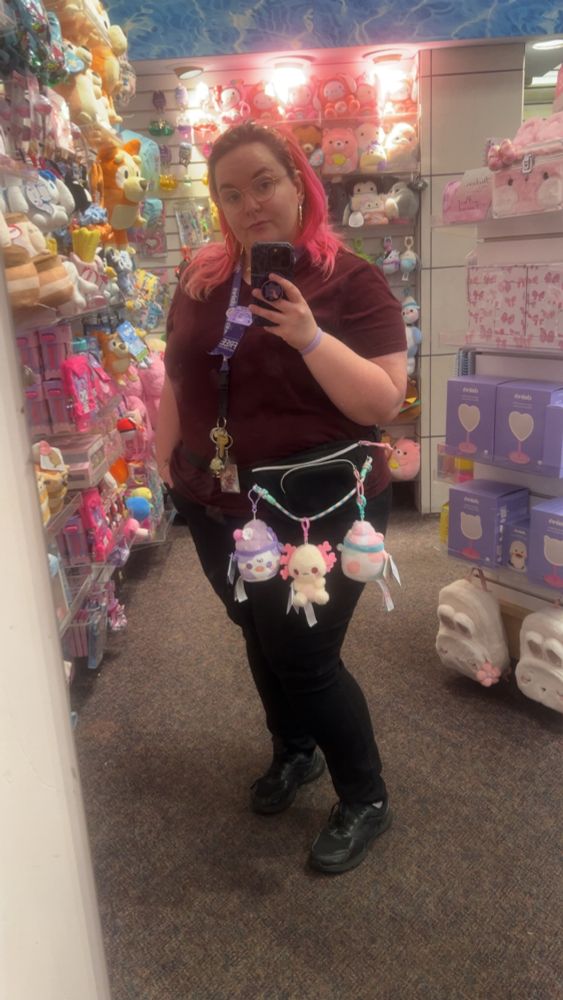 Mirror selfie of pink haired girl with glasses  she’s wearing a Fanny pack with stuffed animals clipped to it 