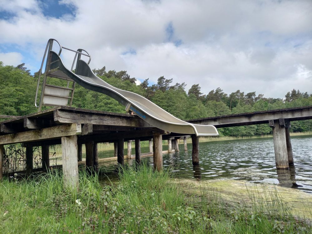Eine gesperrte metallenen Rutsche auf dem Badesteg. Unter dem Ende der Rutsch sind es über 1 Meter zur Wasseroberfläche. Wassertiefe wenige Zentimeter.