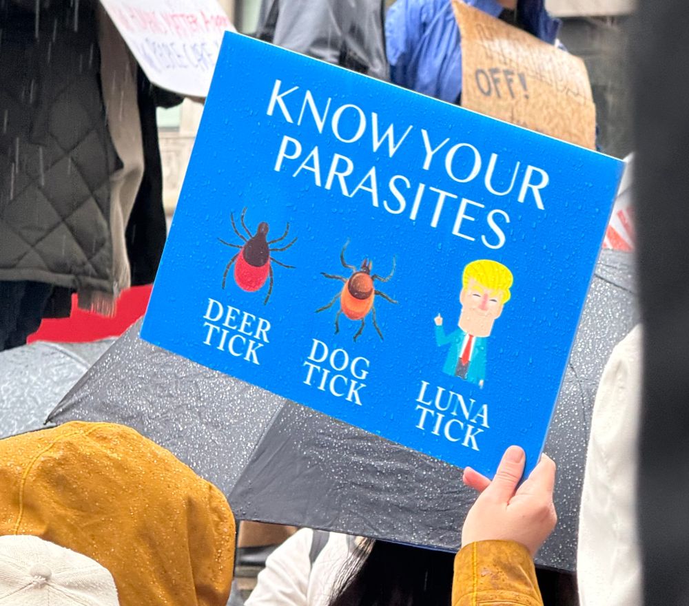 Sign held up at a protest. Sign reads: "Know Your Parasites" underneath, three images, each labeled. First is an image of a tick and it's labeled "Deer Tick." Second is a different kind of tick and it's labelled "Dog Tick." Third is an image of #47 and it is labeled "Luna Tick"