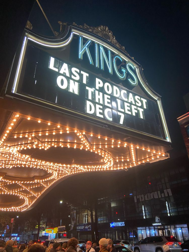 A photo of the Kings Theater awning advertising the Last Podcast on the Left live show December 7. It is lit up festively and crowds meander below. 