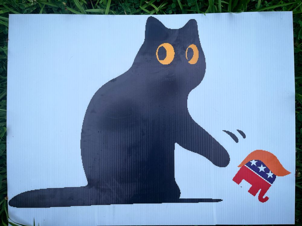 Sign showing a black cat knocking a republicans elephant in a orange wig off a table. 