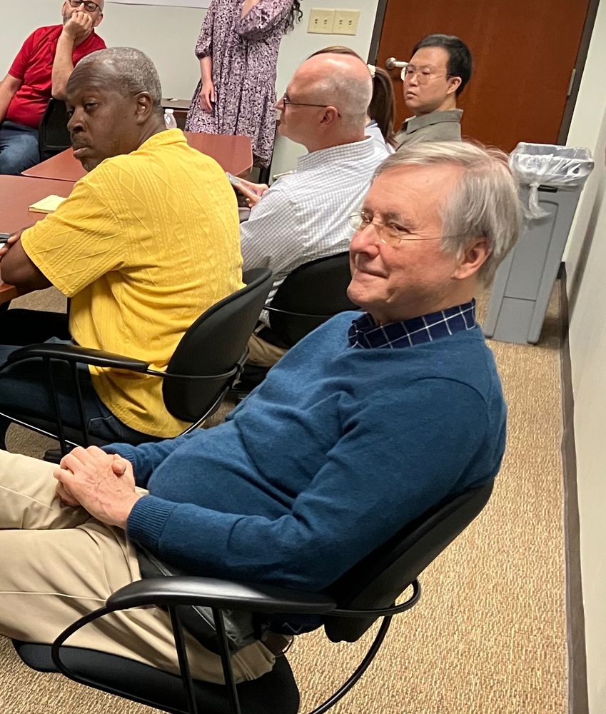 Man with gray hair and wire glasses in blue sweater, blue/white checkered button-down, and khaki pants smiles at someone speaking off-camera.