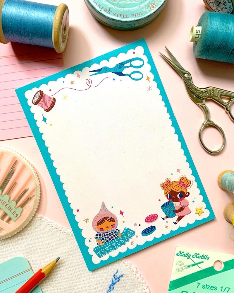 A craft themed illustrated notepad sits on a pink background with a collection of threads, scissors, needles and other craft items strewn about it. The pad itself features illustrated scissors, thread buttons and two little craft inclined characters, one perusing a giant piece of lace and the other holding a huge (for them) thimble.