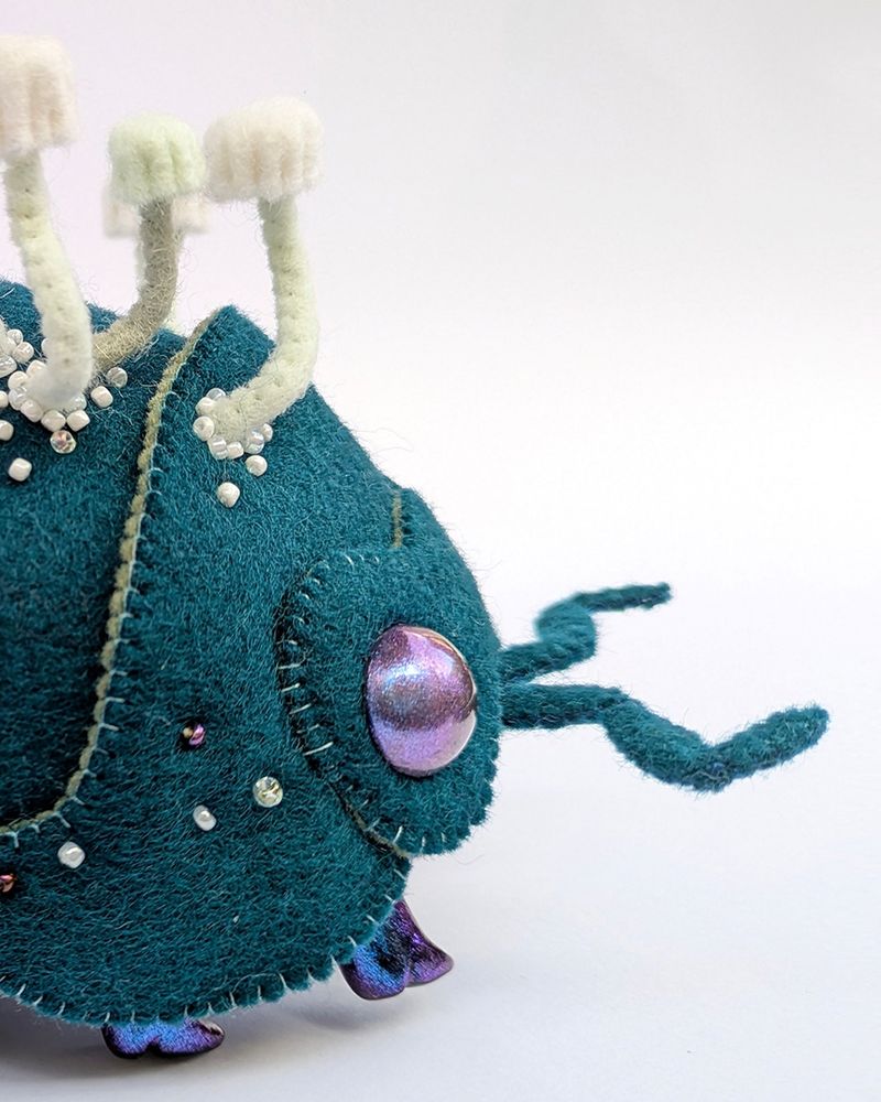 The front half of a side on handmade felt soft sculpture art doll on a white background resembling an isopod/roly-poly bug with plating layers in dark peacock blue on its back and small white mushrooms growing in clusters from it with clusters of mostly white beadwork around them. It has strange purple sparkling eyes and is wearing 10 pairs of hard, sculpted, slightly-heeled sparkling blue/black boots.