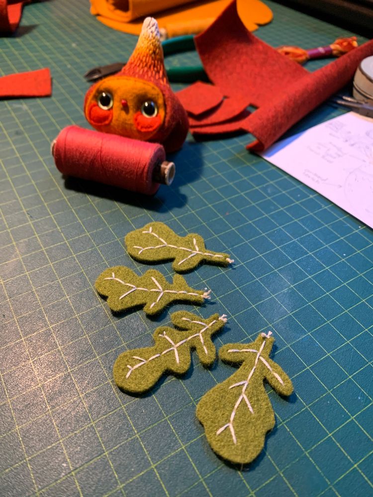 a photo of Hanna’s desk which has a dark green gridded cutting mat with four loose crinkled felt leaves with white embroidered veins sitting on it. Out of focus in the background is some red thread, red and orange pieces of felt and a slightly hard to see handmade red felt bulb/radish with a little face on it.