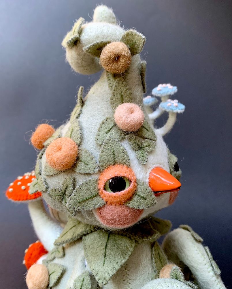 a cropped photo of a hand sewn felt doll with a soft and sage green and orange palette. It has a bulb-ish head, a hard beak, tired looking little eyes and is covered in hand stitched leaves, mushrooms and felt ball flower/berries.