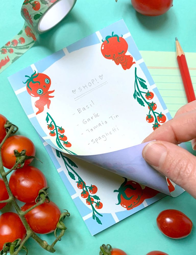 A white hand is half lifting up the top page of a notepad which features anthropomorphic tomato characters and tomato vines with a mint border and cream centre. The pad sits on a pastel green background with real cherry tomatoes strewn about, as well as a red pencil and a tomato themed washi tape.