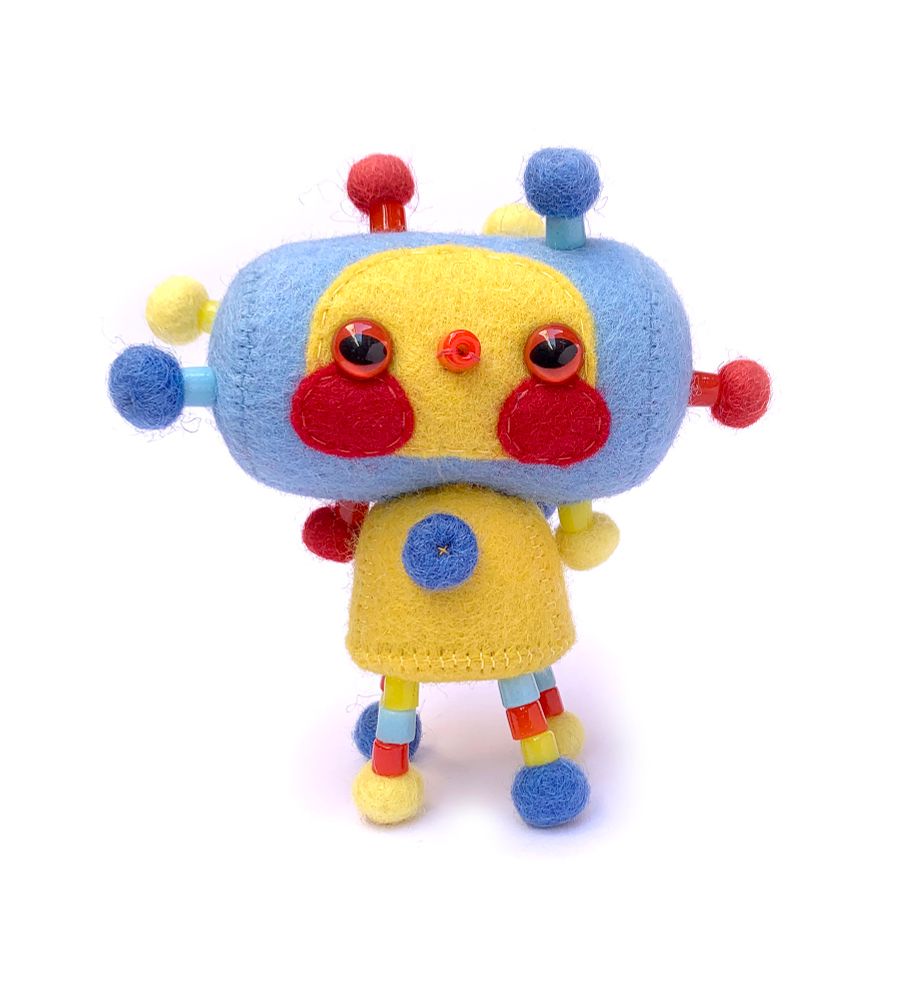 A front on photo of a cute strange handmade felt soft sculpture art doll in primary red, blue and yellow on a whit background. It has a sideways cylindrical blue head with a yellow face, and rosy red cheeks, red eyes and a red mouth made out of a glass bead. It has a small yellow body, 4 skinny beaded legs with pompom feet, and it’s covered all over with yellow, red and blue bead and pompom decorative attachments.