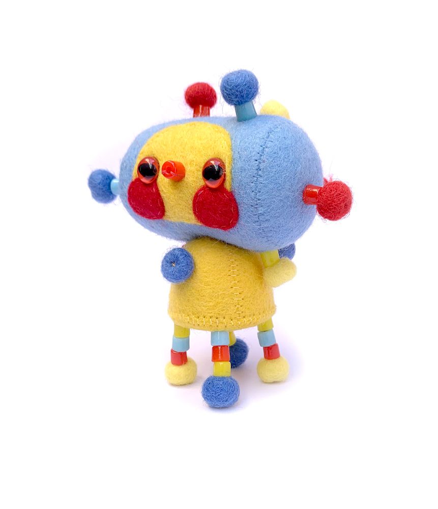 A slightly side on photo of a cute strange handmade felt soft sculpture art doll in primary red, blue and yellow on a whit background. It has a sideways cylindrical blue head with a yellow face, and rosy red cheeks, red eyes and a red mouth made out of a glass bead. It has a small yellow body, 4 skinny beaded legs with pompom feet, and it’s covered all over with yellow, red and blue bead and pompom decorative attachments.