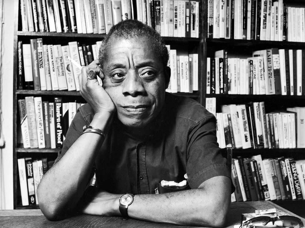 Black and white portrait of James Baldwin sitting before a bookshelf with his cheek resting on his palm, gazing off somewhere. Photo by Ted Thai for LIFE