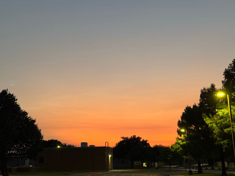A sunset sky that goes from a deep dusky blue at the top to a bright orange at the horizon. A small building and trees are in silhouette along the bottom and lower half of the sides.