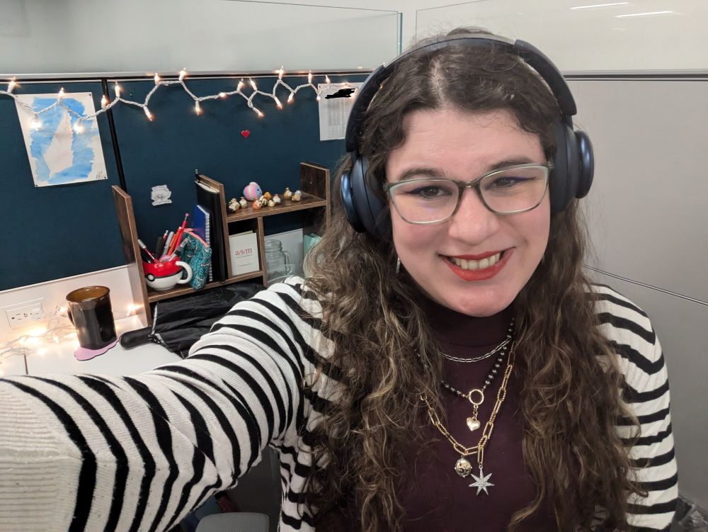 Selfie photo of a woman in a purple sweater dress and striped black-and-white cardigan in an office. Behind her is a desk with christmas lights lining the wall and a small nativity scene on top of a free-standing bookshelf.