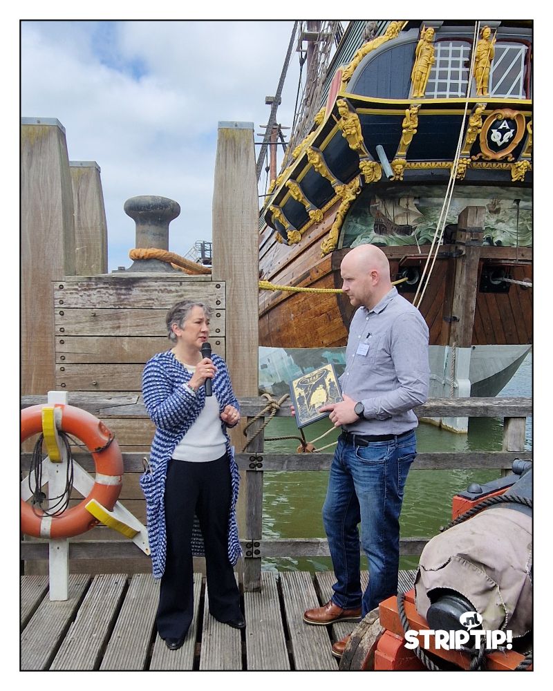 Een foto van de presentatie van het eerste deel van het tweeluik '1629', over de Batavia. De directrice van Museum Batavialand en Dieter Van Tilburgh van Standaard Uitgeverij staan op een steiger, met op de achtergrond de replica van de Batavia. Dieter heeft een exemplaar van de strip in zijn handen. 
