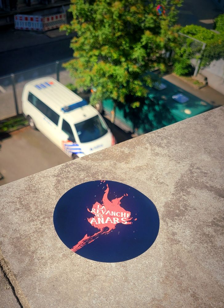 Photo d'un sticker "La Revanche des anars" sur un muret surplombant un combi de flics. Légende : habemus pittacia. En latin, nous avons une étiquette (sticker). Enfin, j'espère que c'est la bonne traduction, hein.