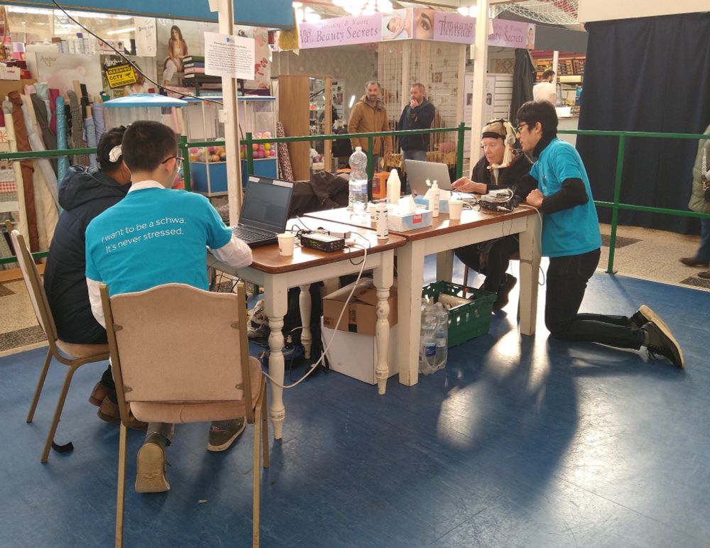 Two people in blue t-shirts are at opposite ends of a table recording ultrasound with two members of the public.