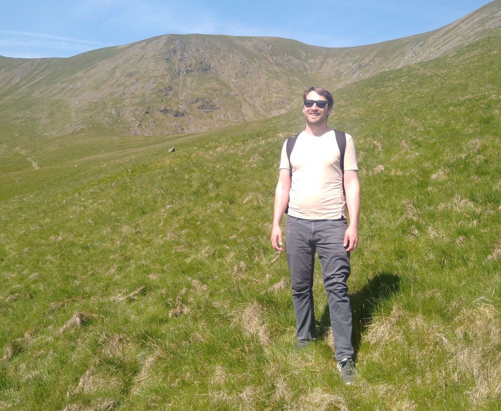 A man is standing on a green grassy mountainside. He is wearing a beige t-shirt, sunglasses, and a rucksack. The sky is blue and the grass is very green.