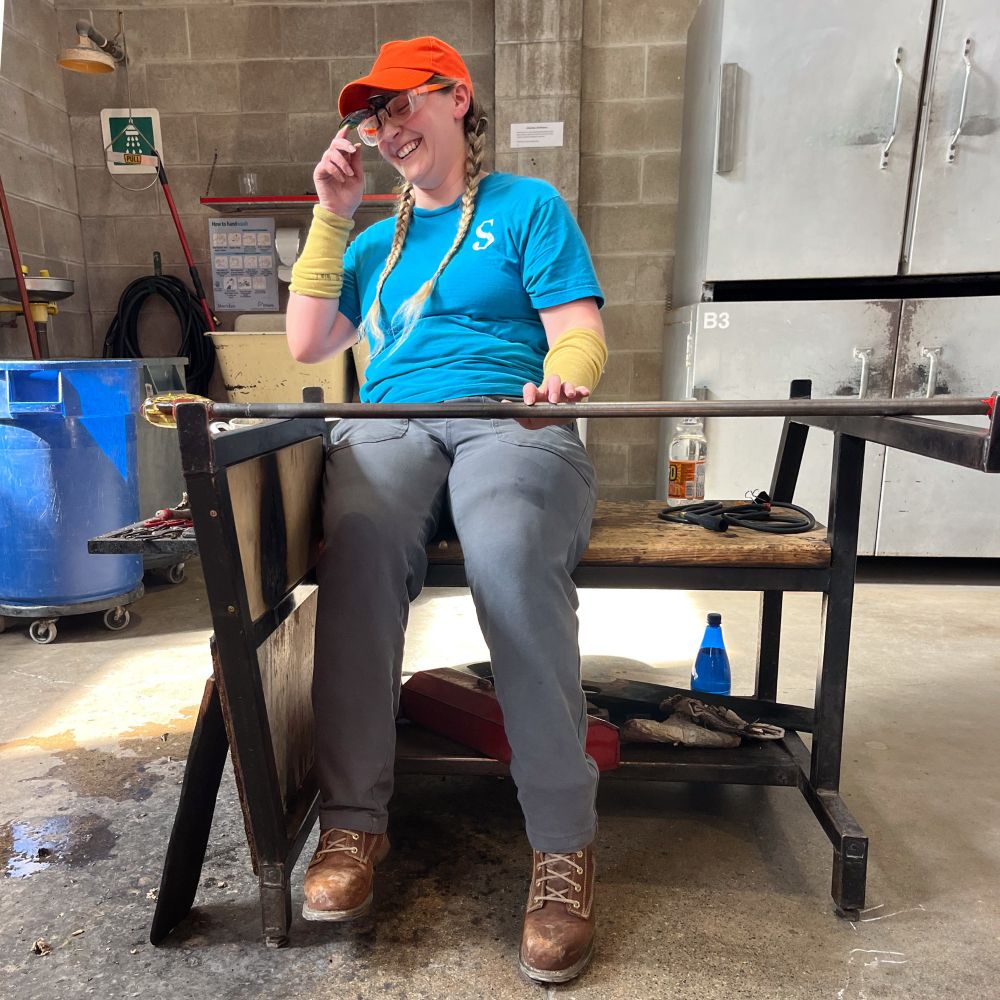 Photo of a smiling person blowing glass in a hot glass studio 