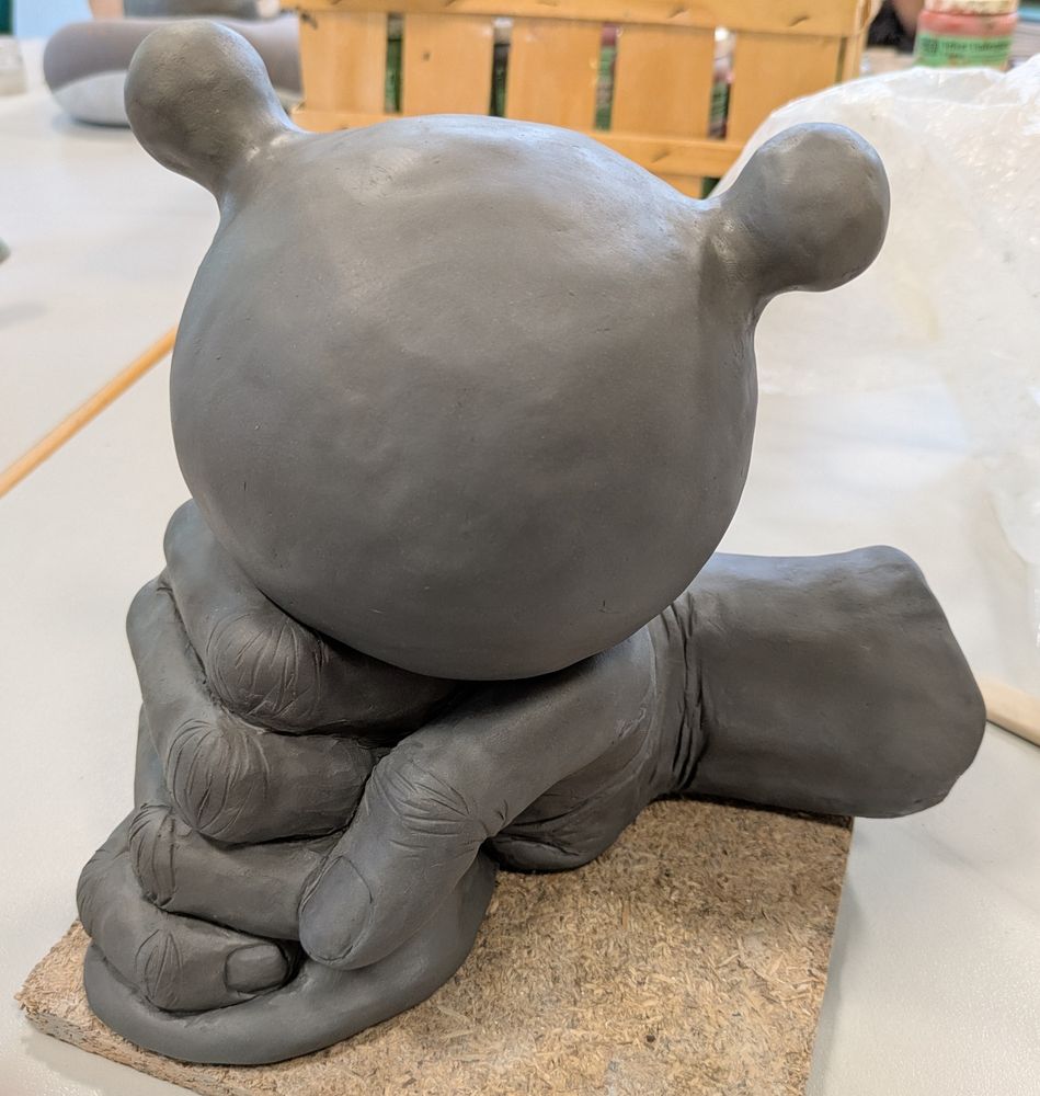 Fresh clay sculpture of a giant muscular fist squeezing anti stress ball. 2 smaller balls (eyes?) protrude from the anti stress ball. Bottom of the anti stress ball is pressed flat on the table.