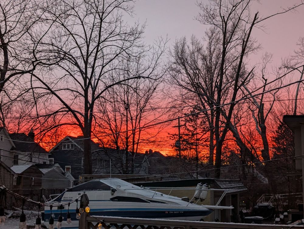 A sunset image. At the top the sky is a blue gray, but becomes pale pink, and finally a brilliant orange. In the foreground are trees, houses, and a boat (in my neighbor's yard, not in water), everything under a dusting of new snow 