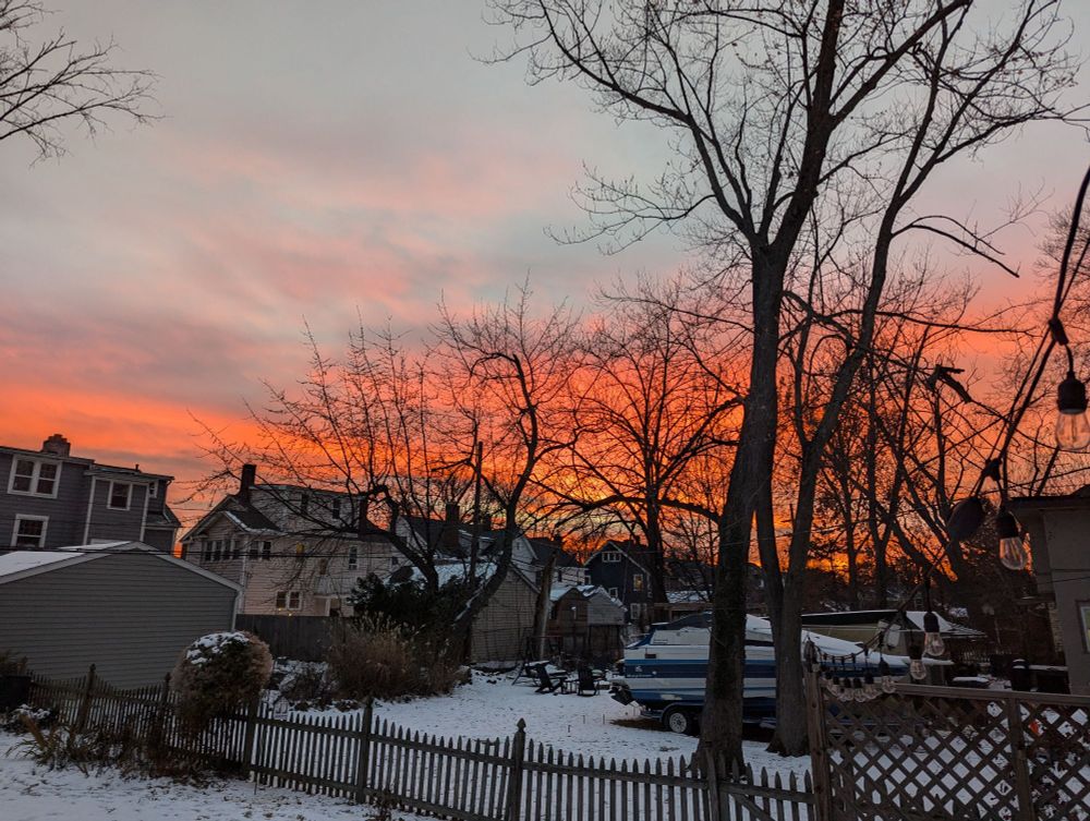 A winter landscape. Snow on the ground, bare trees in the middle distance and a sky that has blue sky with clouds but lower down the sky goes from pink to orange to a fiery orange 