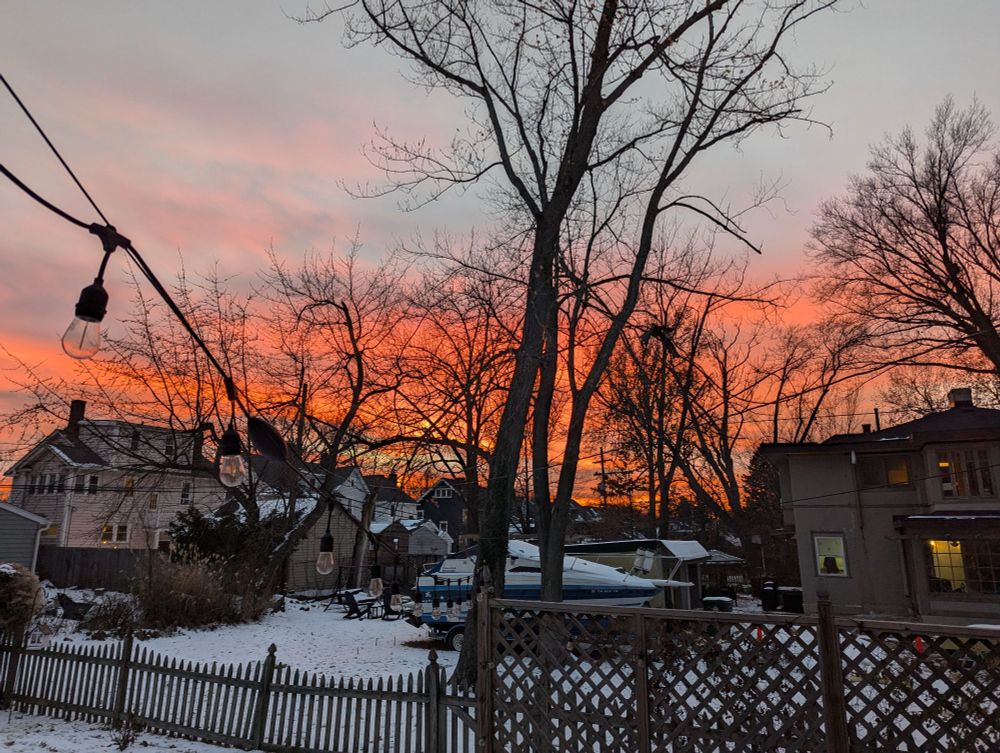 A winter landscape. Snow on the ground, bare trees in the middle distance and a sky that has blue sky with clouds but lower down the sky goes from pink to orange to a fiery orange 