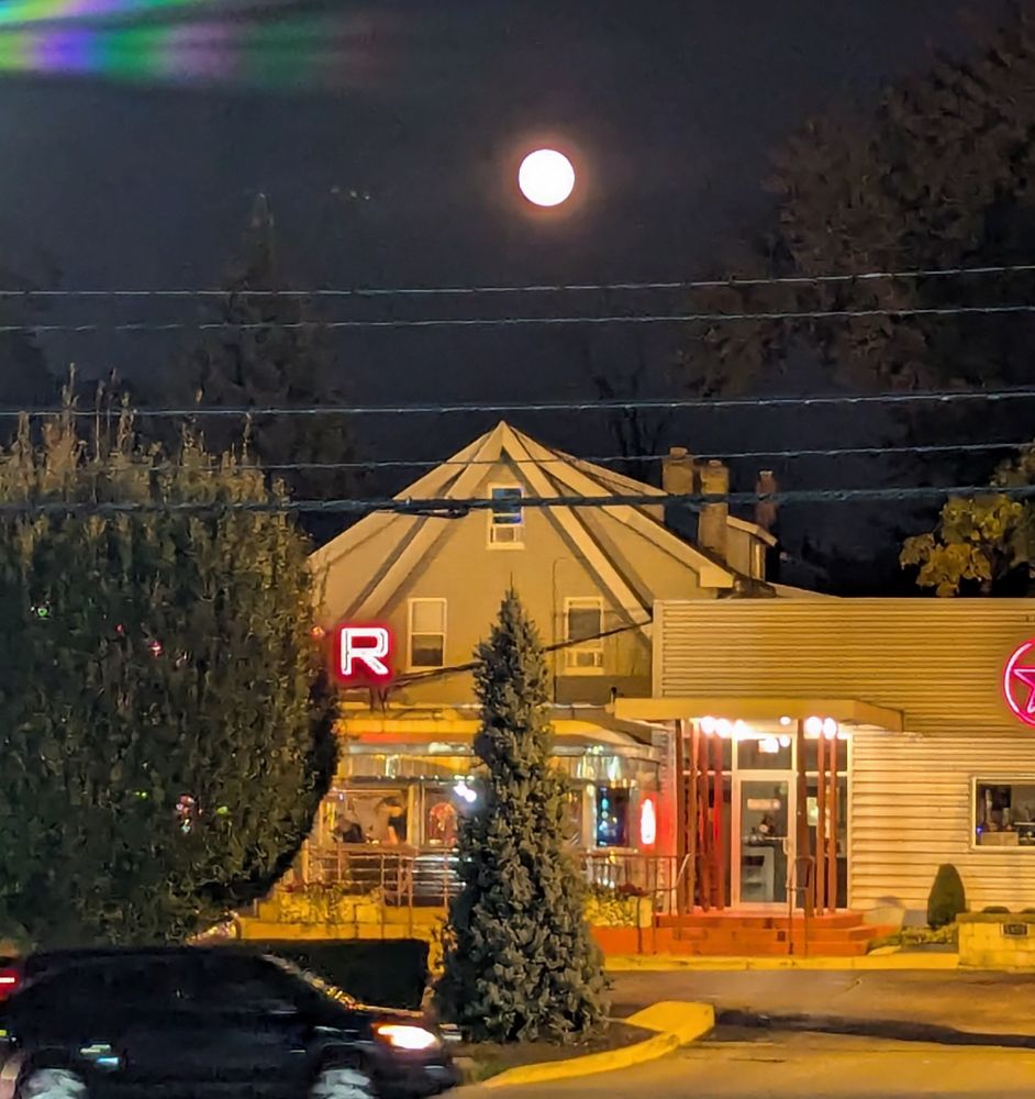 Full moon rising above a house and a diner, trees and a passing car in the foreground 