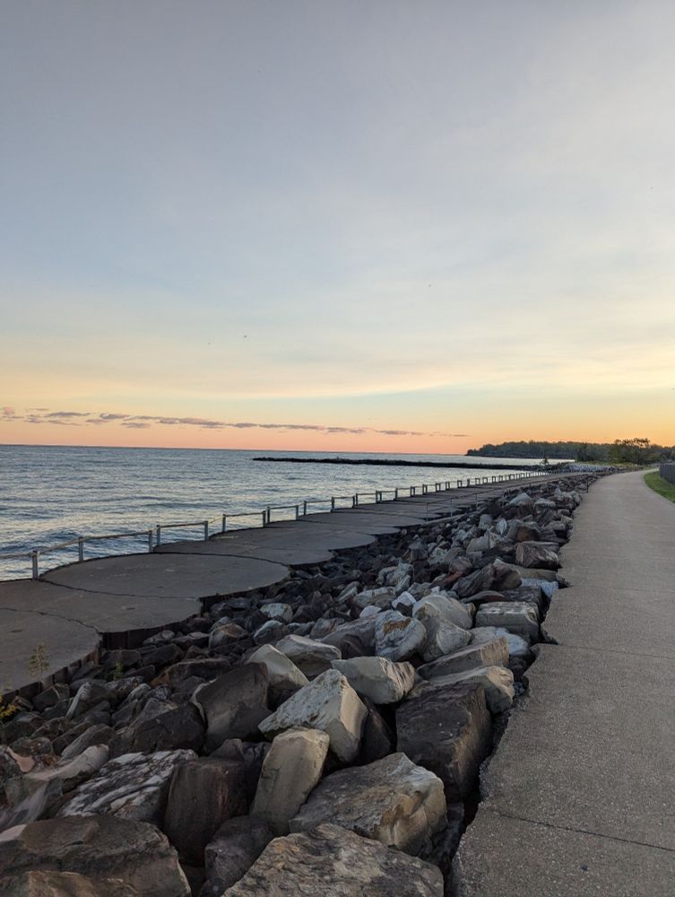A light pink and blue sky over a big body of water, rocks and paths in the foreground.