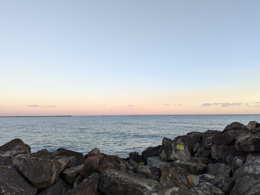 A light pink and blue sky with wispy clouds over a lake as the sun rises. In the foreground are large rocks.