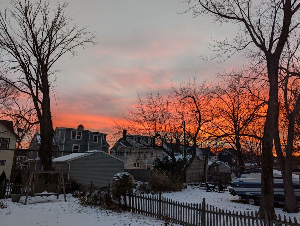 A winter landscape. Snow on the ground, bare trees in the middle distance and a sky that has blue sky with clouds but lower down the sky goes from pink to orange to a fiery orange 
