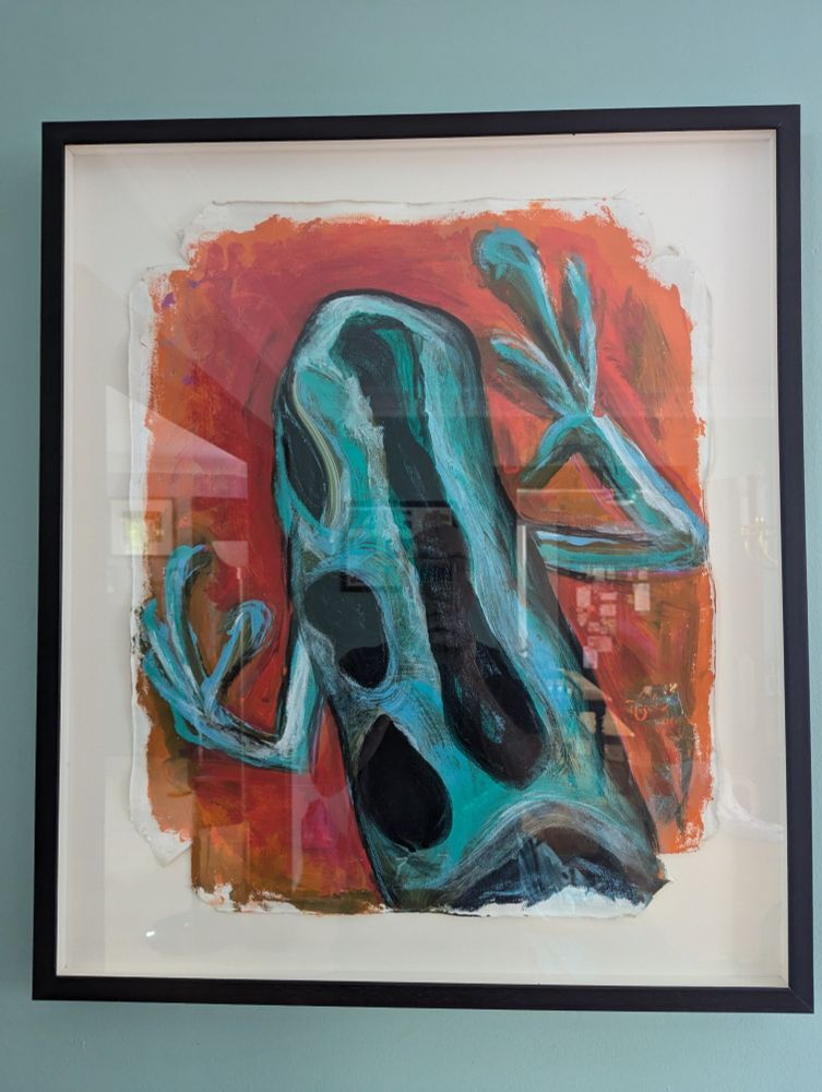 A framed painting of a frog. The frog is blue-green with black spots. The background of the painting is shades of red and orange.