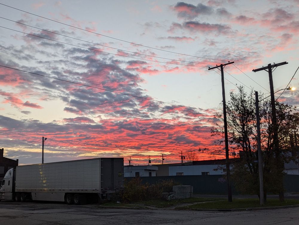 An early morning sky with wispy pink clouds in the foreground and fiery red clouds in the background. There are trucks and utility poles below.