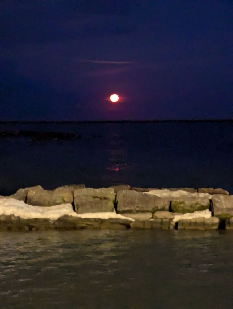 The moon, looking like a small orange orb, setting over Lake Erie. The sky behind the moon is dark blue with some light wispy clouds. Below the lake is almost black. In the foreground are rocks from the break wall and more water.