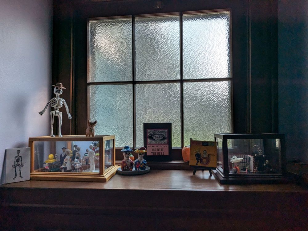 Many pieces of Day of the Dead art. Some are in glass display cases, all or on a wooden mantle. There is a window in background that has pebbled glass giving a blur effect to the light coming through.